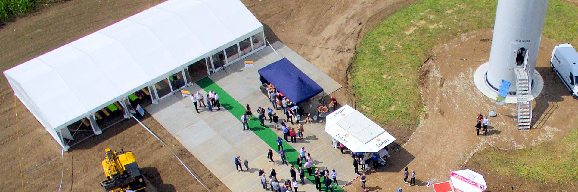 Zu sehen ist ein Luftbild mit Blick auf den Fuß einer Windenergieanlage mit einem Festzelt, Menschen und Imbissständen.